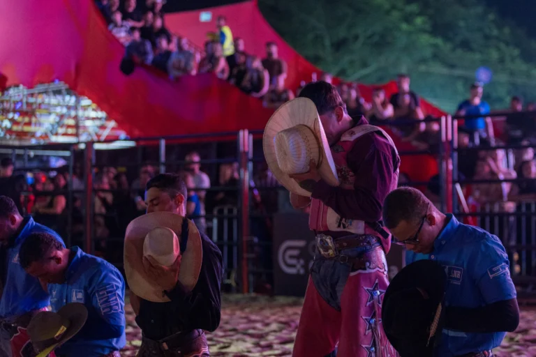 Peões ajoelhados na arena durante a abertura do Limeira Rodeo Music com a imagem de Nossa Senhora Aparecida e fogos.