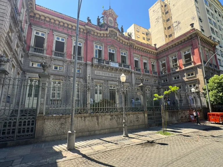 Estudantes protestando na frente de uma escola, com cartazes denunciando assédio e policiais ao fundo.