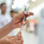 Foto de braço recebendo a vacina da gripe, com pessoas ao fundo em uma UBS de Araçatuba