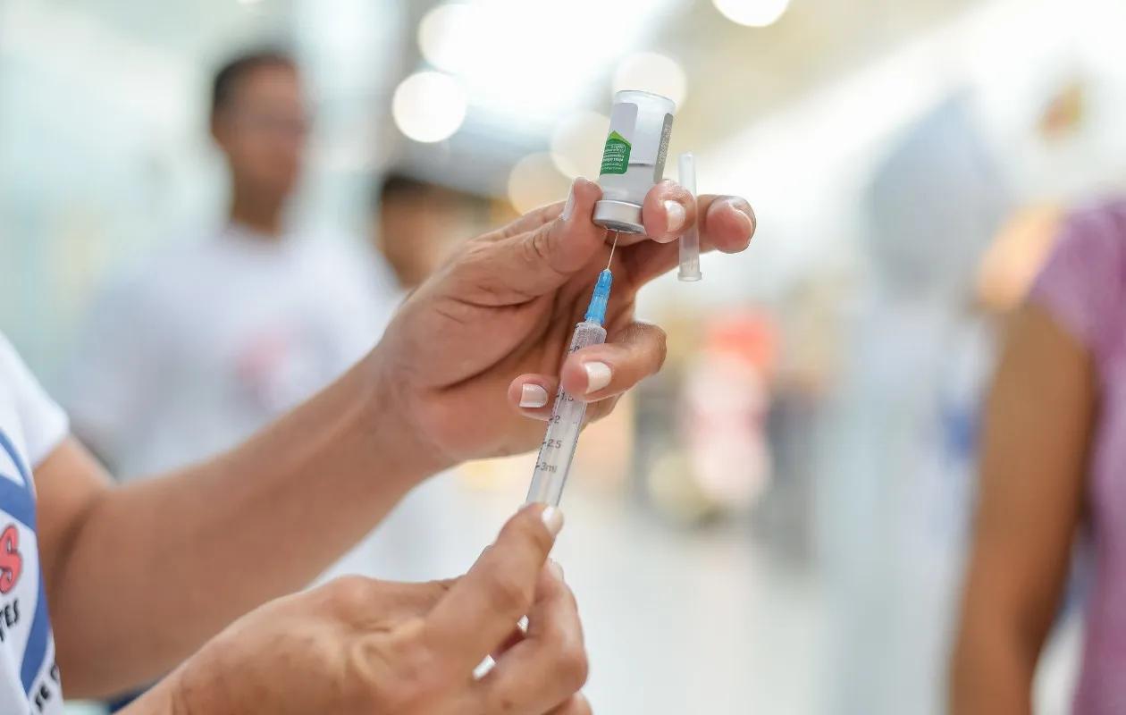 Foto de braço recebendo a vacina da gripe, com pessoas ao fundo em uma UBS de Araçatuba