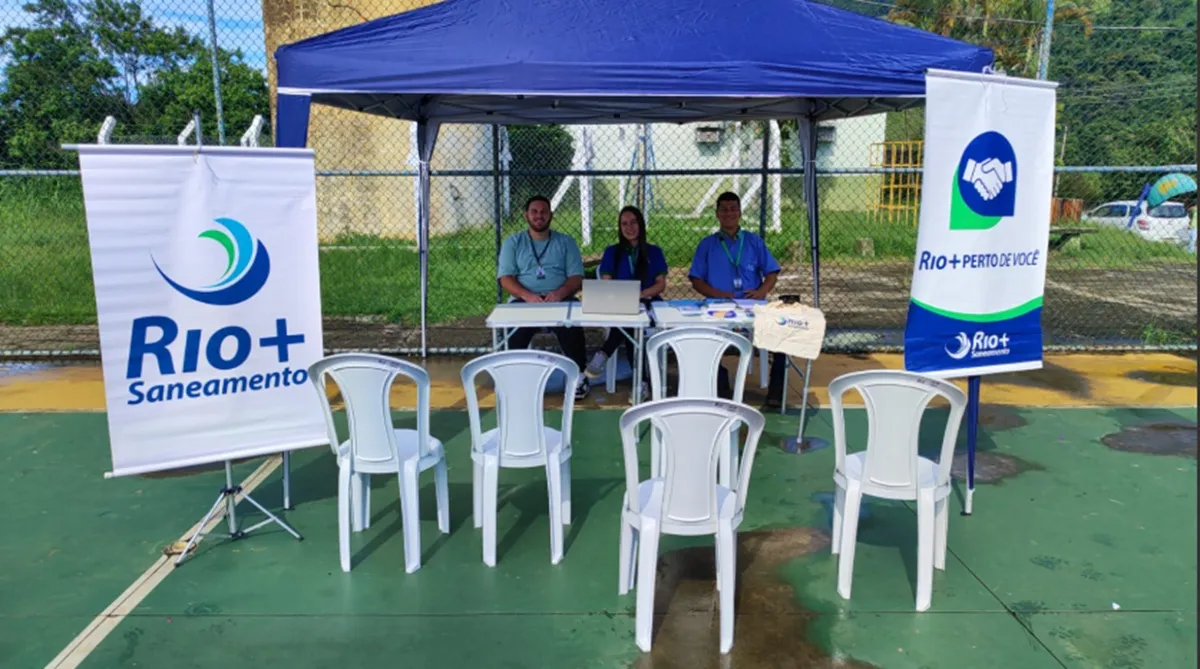 Atendimento itinerante da Rio+Saneamento em praça pública, com moradores sendo atendidos por equipe da concessionária para serviços de água e esgoto.