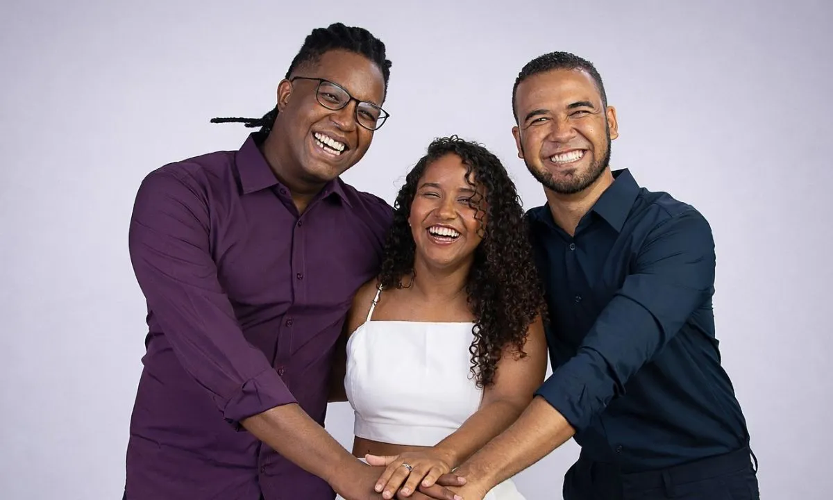 Trio de baianos criadores da ferramenta de IA Yá, sorrindo e celebrando em palco de conferência internacional.
