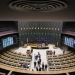 Gavel sobre mesa de madeira, com plenário da Câmara ao fundo, representando a aprovação de leis.