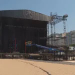Palco do show de Shakira em construção na Praia de Copacabana ao pôr do sol, com equipamentos e guindastes.