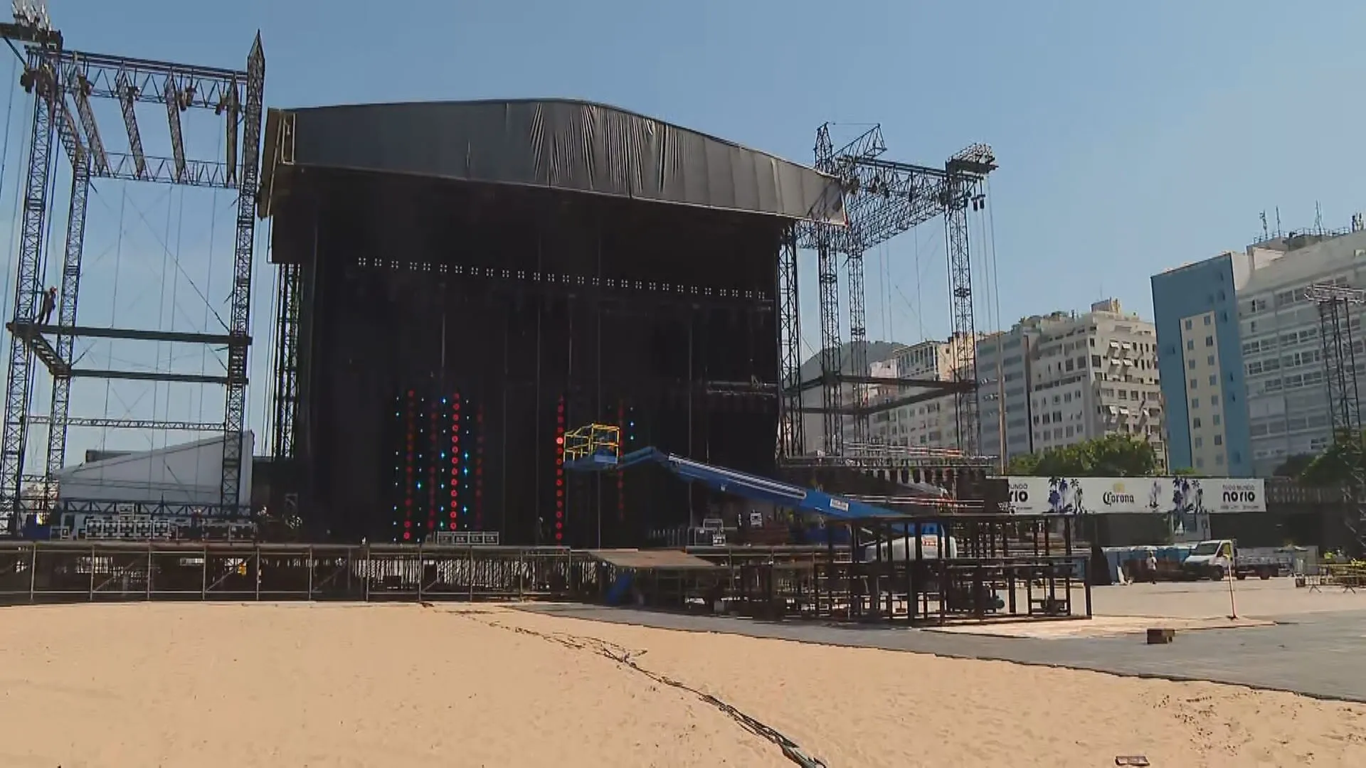 Palco do show de Shakira em construção na Praia de Copacabana ao pôr do sol, com equipamentos e guindastes.