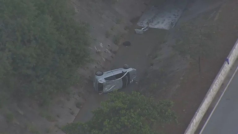 Carro capotado e parcialmente submerso em córrego na Avenida Jacu-Pêssego, com bombeiros e polícia no local.