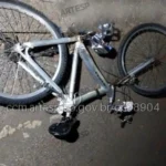 Foto aérea de um acostamento de rodovia à noite, com luzes de emergência e destroços de bicicleta, simbolizando o acidente fatal.