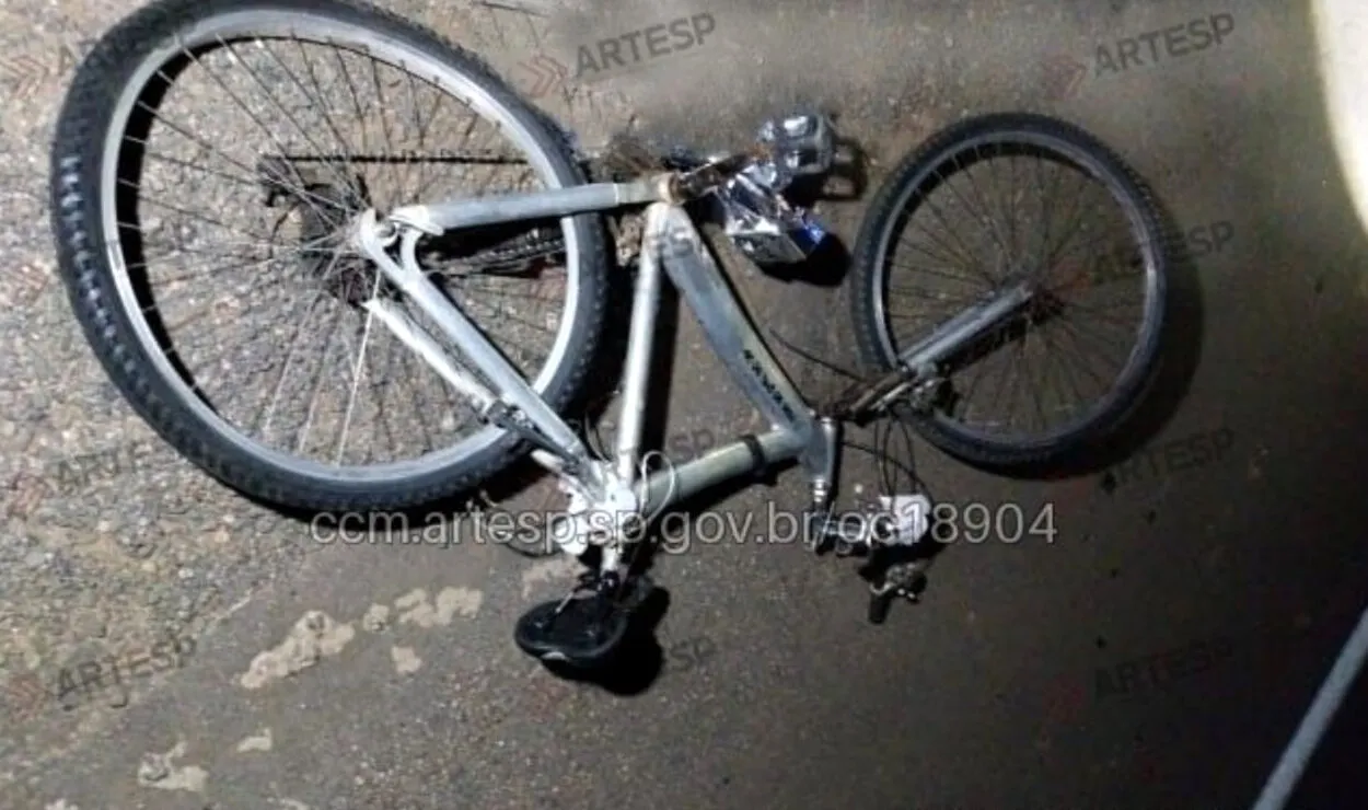 Foto aérea de um acostamento de rodovia à noite, com luzes de emergência e destroços de bicicleta, simbolizando o acidente fatal.