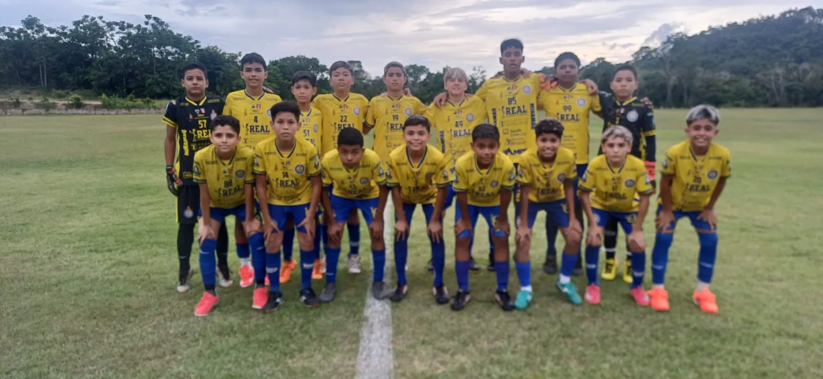 Jogadores de futebol juvenil disputando uma bola em campo de gramado verde, com torcedores ao fundo.