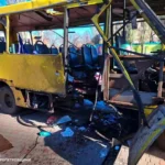 Ônibus civil destruído por drone russo em Nikopol, Ucrânia, com danos visíveis e equipes de resgate avaliando a situação.