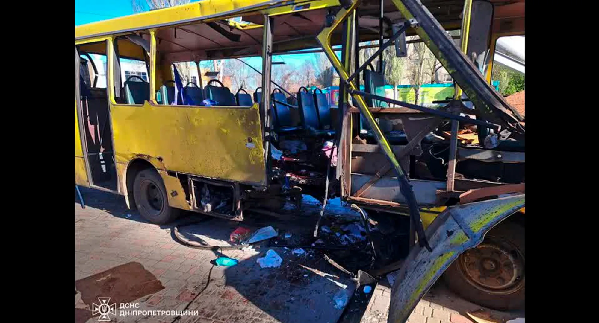 Ônibus civil destruído por drone russo em Nikopol, Ucrânia, com danos visíveis e equipes de resgate avaliando a situação.