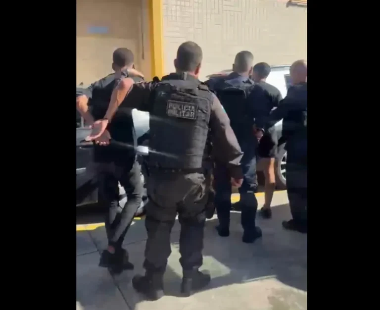 Polícia Militar detendo dois homens, um adulto e um adolescente, após roubo a idosa em Petrópolis, com viatura e rua ao fundo.