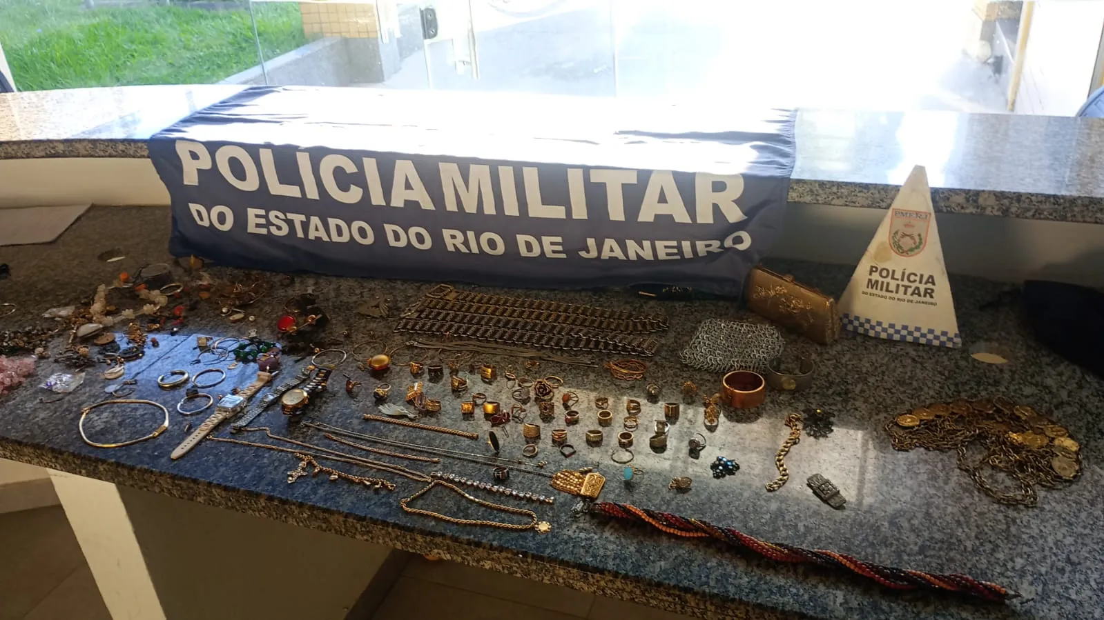 Bolsa aberta cheia de joias (anéis, brincos, pulseiras) sendo inspecionada por um policial.