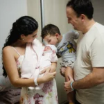 Jovem mãe Maria Eduarda abraça seu bebê recém-nascido após um parto rápido no banheiro de casa, com um olhar de superação e amor.