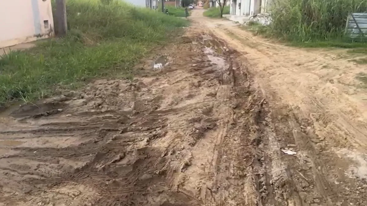 Rua esburacada e enlameada no bairro João Bedim, Itaperuna, com moradores caminhando com dificuldade e carros parados.