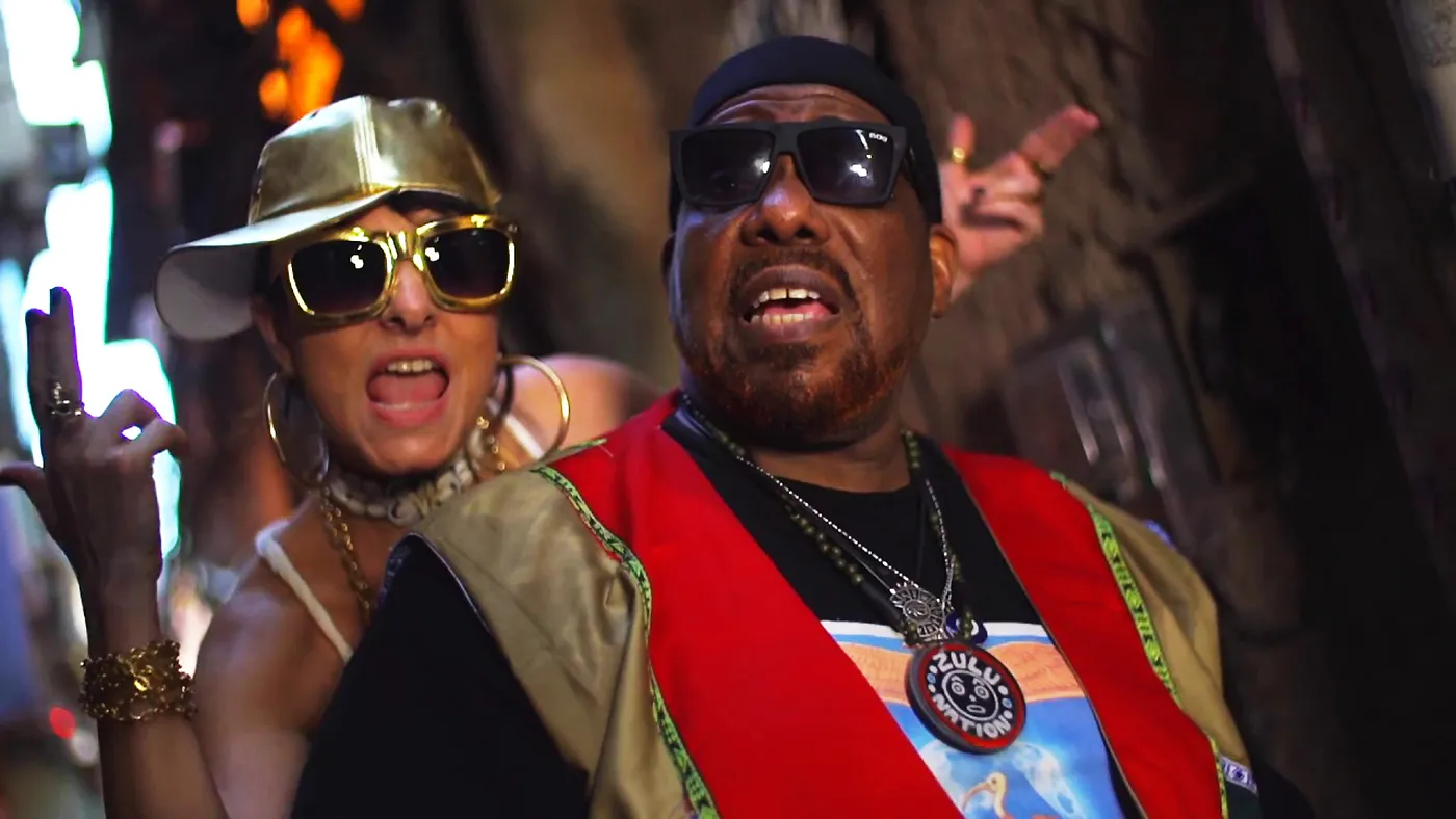Fernanda Abreu e Afrika Bambaataa no set de 'Tambor', com tambores e cenário de favela carioca.