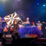 Pessoas assistindo a uma apresentação no Festival de Curitiba, com arquitetura da cidade ao fundo.