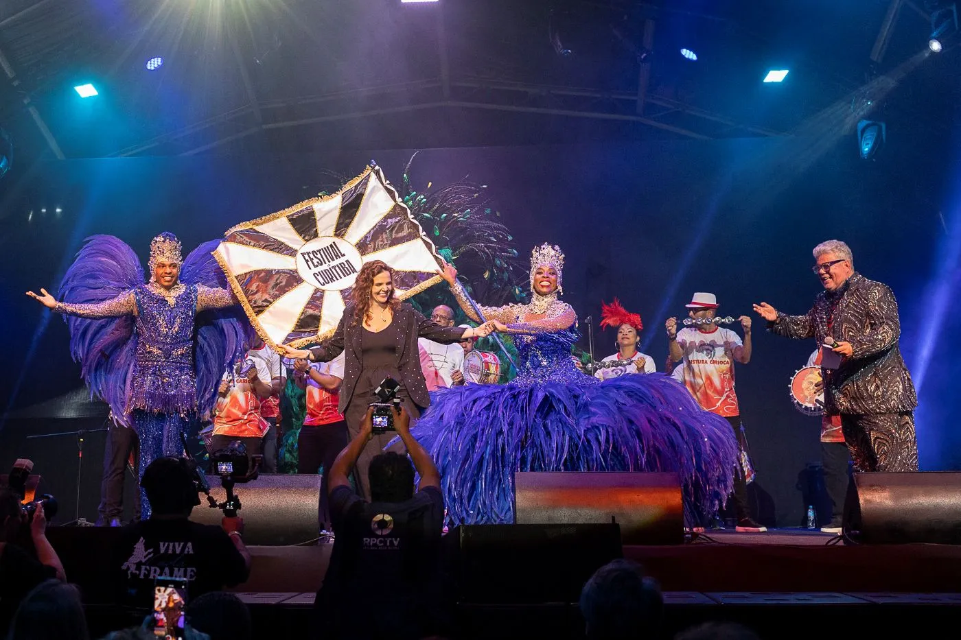Pessoas assistindo a uma apresentação no Festival de Curitiba, com arquitetura da cidade ao fundo.
