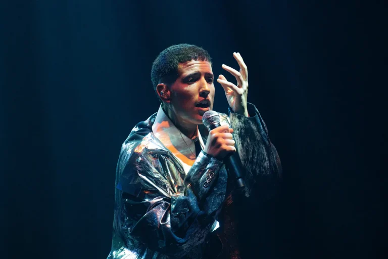 Cantor Fitti no palco do Queremos! Festival, interpretando canções de Ney Matogrosso, com banda ao fundo e iluminação de show.