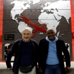 Pierre Guillon de Prince e Dieudonne Boutrin próximos a um mastro de navio em Nantes, simbolizando a reconciliação e o pedido de desculpas pela escravidão.