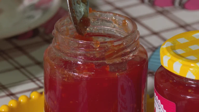 Pote de geleia de tomate com pimenta caseira, tomates frescos e pimentas vermelhas em uma mesa de madeira.