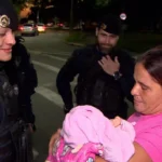 Mãe segurando bebê recém-nascido, com guarda municipal ao fundo, após resgate de engasgo.