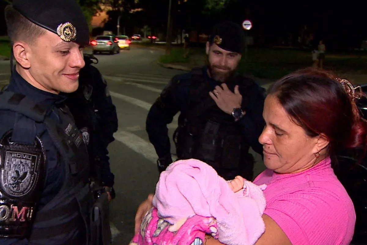 Mãe segurando bebê recém-nascido, com guarda municipal ao fundo, após resgate de engasgo.