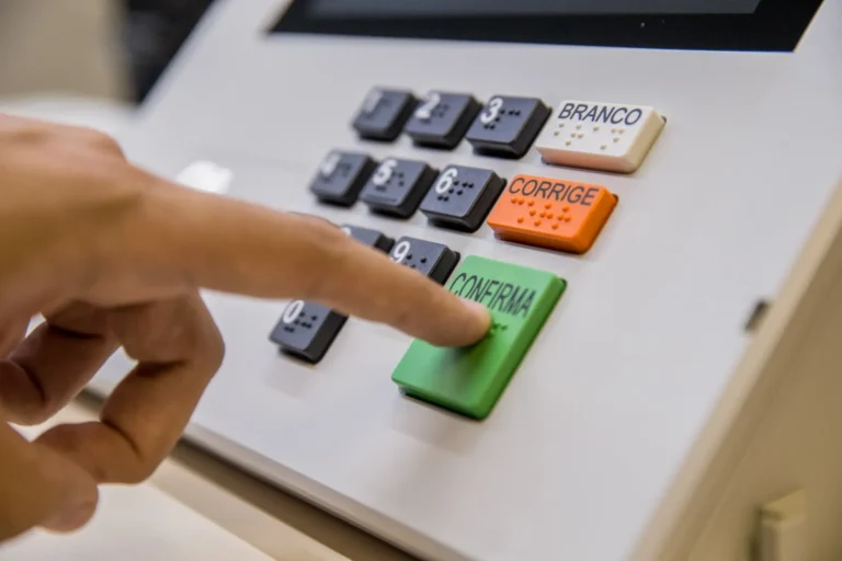 Urna eletrônica com mão de eleitor votando, fundo desfocado de seção eleitoral, eleitores do RJ