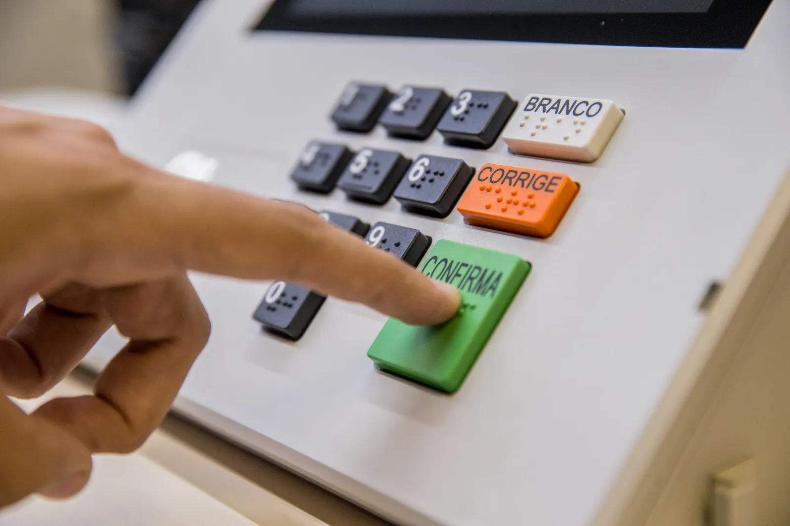 Urna eletrônica com mão de eleitor votando, fundo desfocado de seção eleitoral, eleitores do RJ