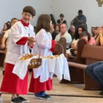 Menino com Síndrome de Down vestindo roupa de coroinha e sorrindo na igreja.