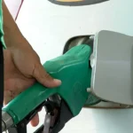 Bico de bomba de combustível de etanol abastecendo carro, com fundo de campos de cana de açúcar, representando a nova política de combustível e o aumento do teor de etanol na gasolina.