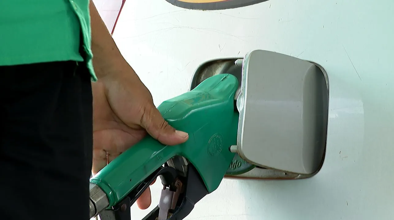 Bico de bomba de combustível de etanol abastecendo carro, com fundo de campos de cana de açúcar, representando a nova política de combustível e o aumento do teor de etanol na gasolina.