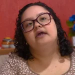 Mulher jovem com deficiência, óculos, sorrindo em ambiente de trabalho, representando inclusão e empoderamento.