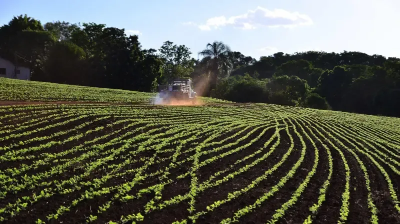 Navio cargueiro no Estreito de Ormuz com campos agrícolas ao fundo, simbolizando a dependência do agronegócio brasileiro de fertilizantes importados e a ameaça da guerra no Irã.