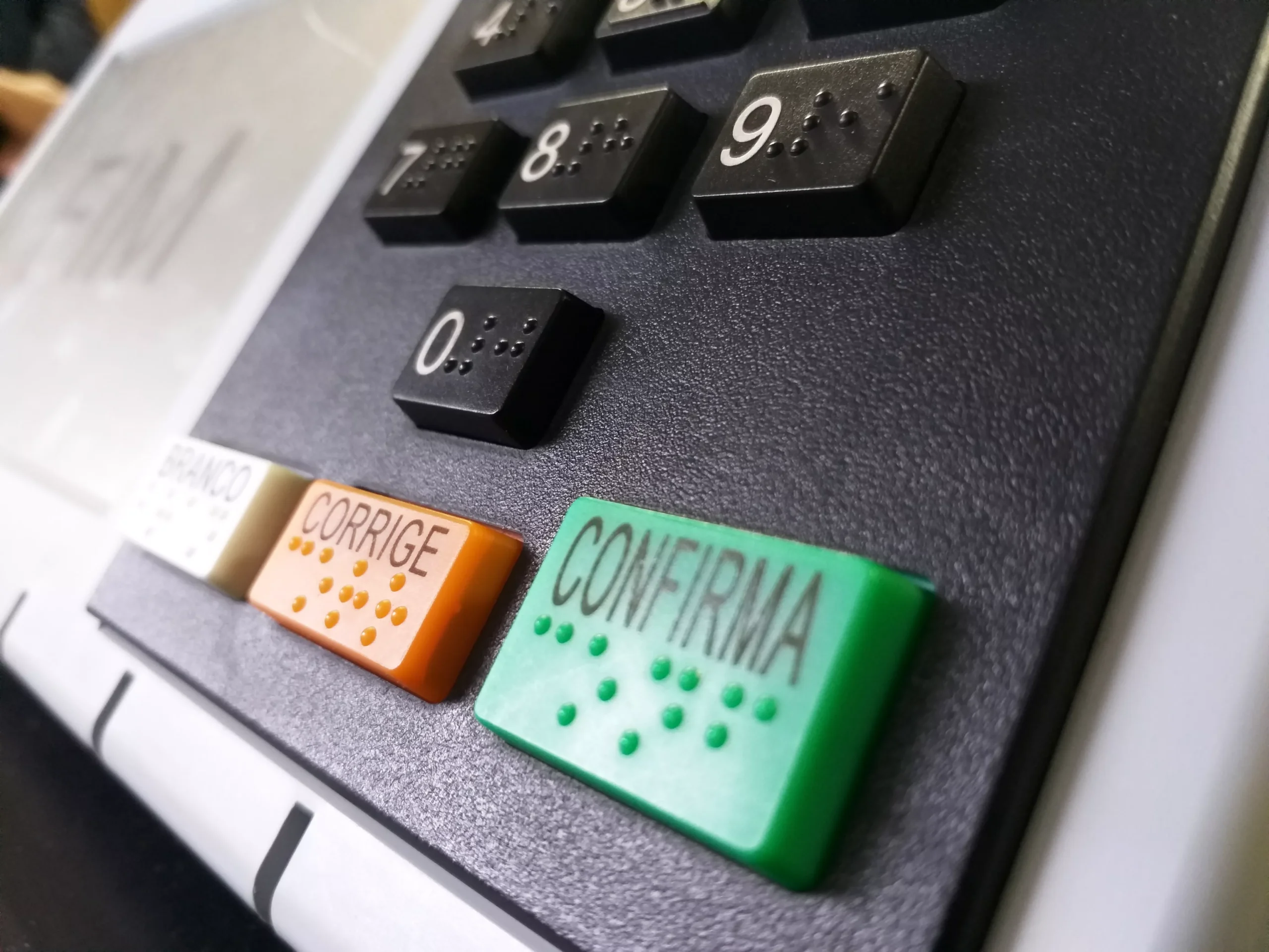 Urna eletrônica recebendo voto, com foco na mão do eleitor e fundo de pessoas diversas