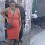 Mulher algemada sendo escoltada por policiais em uma rua de comunidade, representando a prisão por fraude em leilões de carros.