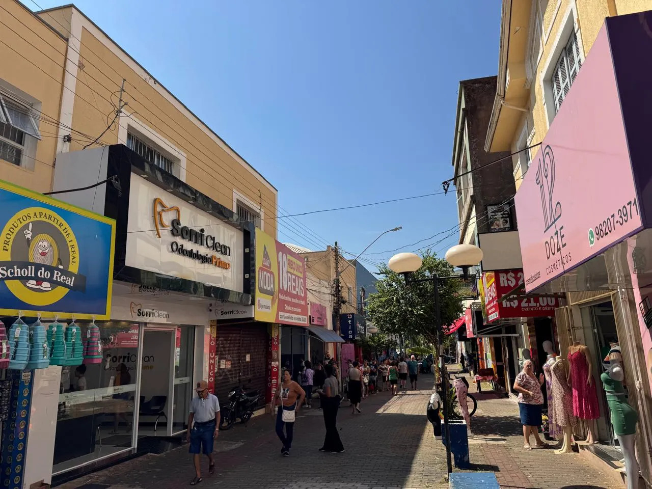 Pessoas em rua comercial movimentada durante feriado de Tiradentes, com lojas abertas e movimento de pedestres.
