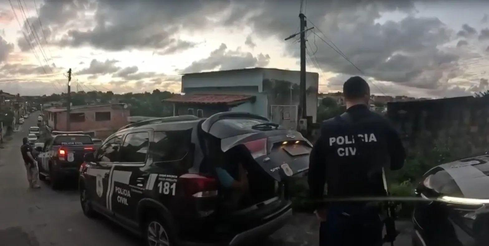 Três homens algemados sendo escoltados por policiais, com celulares e uma moto apreendidos em segundo plano, representando a prisão por roubo de celulares.