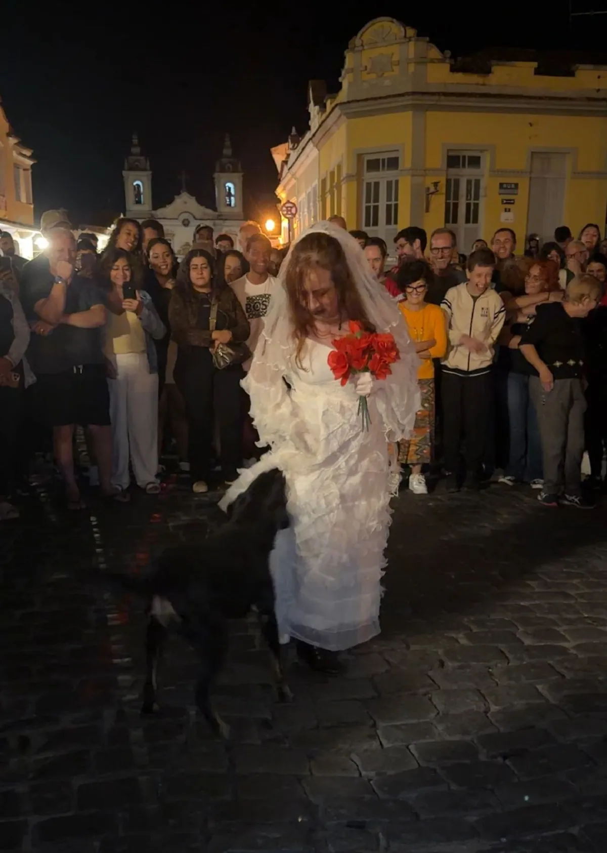 Vira-lata no teatro de rua, puxando vestido de atriz durante peça dramática.